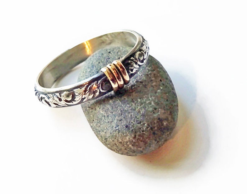 May include: A silver ring with an engraved floral pattern and gold accents. The ring is resting on a gray stone against a white background. The ring's design features intricate details and a contrasting color scheme.