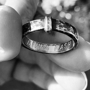 May include: Close-up of a silver ring held between fingers. The ring has a rectangular accent and the engraved words "DINNA FASH" on the inside. The ring appears to be worn and has a vintage aesthetic.