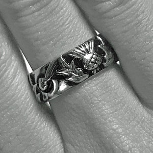 May include: A silver ring with a floral design. The ring features detailed carvings of flowers and leaves around the band. The ring is worn on a finger.