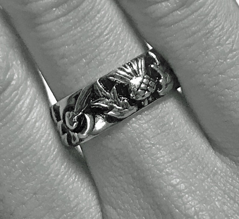 May include: A silver ring with a floral design. The ring features detailed carvings of flowers and leaves around the band. The ring is worn on a finger.
