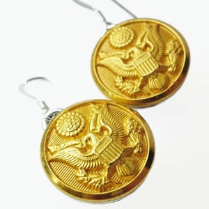 May include: A pair of gold-colored button earrings with silver-colored hooks. Each earring features a detailed embossed design of an eagle, shield, and other symbols. The earrings are round and have a shiny finish.