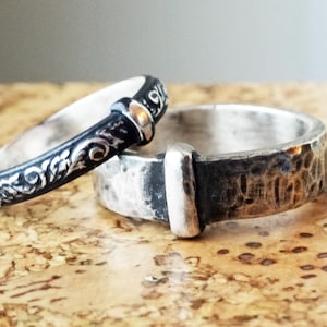 May include: Two silver rings. One ring is black with a floral design and a silver accent. The other ring is silver with a hammered texture and a silver accent. The rings are on a brown surface.