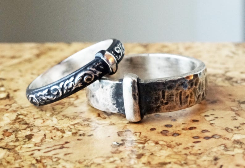 May include: Two silver rings. One ring is black with a floral design and a silver accent. The other ring is silver with a hammered texture and a silver accent. The rings are on a brown surface.