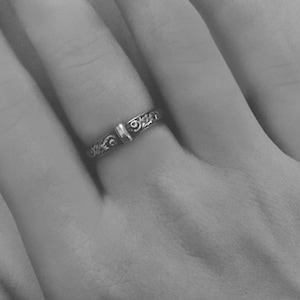 May include: A silver ring with an ornate, swirling design is worn on a finger. The ring has a central, raised element and detailed patterns along its band. The image is in black and white.