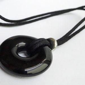 May include: A black, donut-shaped pendant necklace. The pendant is strung on a black cord with a silver-coloured metal accent. The necklace is shown against a white background.