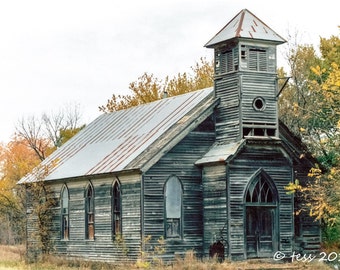 May include: A weathered, gray wooden church with a steeple and a rusty metal roof. The church has several windows and a large, arched doorway. The church is surrounded by trees with fall foliage.