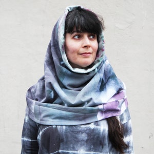 May include: A tie-dye hooded scarf in shades of blue, purple, and gray. The scarf is draped around the neck and over the head, creating a layered effect. The fabric appears soft and comfortable, with a subtle pattern throughout.