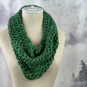 May include: A green crocheted infinity scarf displayed on a white mannequin. The scarf is crafted using a basic stitch pattern, giving it a subtly textured appearance.