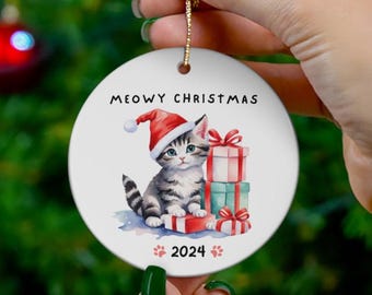 May include: A white ceramic Christmas ornament with a cartoon illustration of a gray and white kitten wearing a red Santa hat and holding a stack of wrapped gifts. The ornament reads "MEOWY CHRISTMAS 2024".