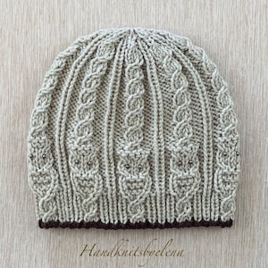 Peut inclure: Un bonnet en tricot beige avec un motif en câble et une bordure marron. Le bonnet est décoré de petites perles argentées en forme de hiboux.
