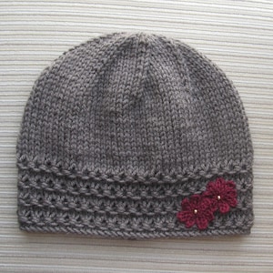 Könnte beinhalten: Grau gestrickte Mütze. Die Mütze hat ein dekoratives Band mit einem strukturierten Muster und zwei kleinen, dunkelroten Blumen mit goldfarbenen Zentren. Diese Mütze ist für Wärme und Stil konzipiert.
