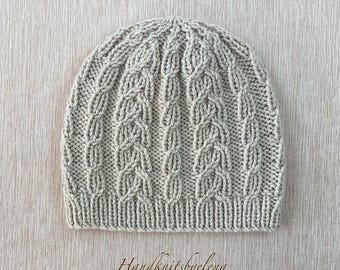 May include: A light beige knit beanie featuring a vertical cable knit pattern throughout the body and a ribbed knit brim at the base. The text Handknitsbyelena is printed in a cursive script font below the beanie on the surface.