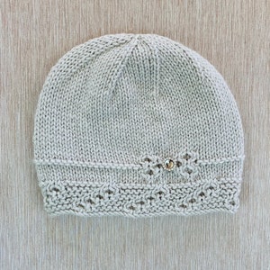 Puede incluir: Un gorro de punto beige con un delicado patrón de encaje alrededor del borde y un pequeño botón brillante en la parte superior.