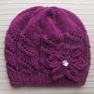 Puede incluir: Un gorro tejido de color magenta con un diseño floral y un centro de pedrería. El gorro presenta un patrón texturizado y una flor decorativa en el lateral. Este gorro hecho a mano es un accesorio elegante para el clima frío.