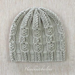 May include: A light grey knitted beanie hat with a textured cable knit design. The hat has a ribbed brim and vertical cable patterns. The text "Handknitsbyelena" is written in cursive at the bottom of the image.