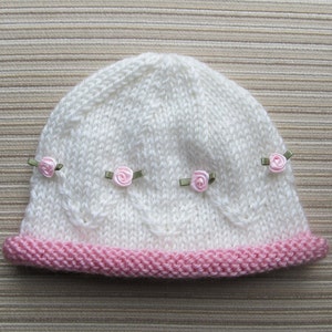 May include: A white knitted beanie featuring a pink garter stitch brim and a series of small pink ribbon roses with green leaves attached in a horizontal row across the center, accented by a subtle cable knit pattern integrated into the white yarn.