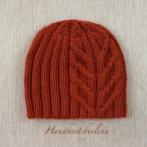 May include: A rust-colored knit beanie featuring a vertical ribbed pattern on the left side and a prominent, textured cable knit design on the right side. The hat is crafted from yarn and displays the text Handknitsbyelena at the bottom center.