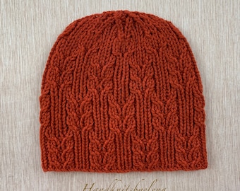 May include: A rust-coloured knitted beanie featuring a repeating vertical cable stitch pattern that resembles stalks of wheat. The hat is constructed with a thick, textured yarn and displays a consistent, dense knit structure throughout the entire piece.