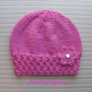 May include: A bright pink knitted beanie featuring a textured cable knit pattern along the bottom edge and a crocheted flower embellishment with a clear, faceted center stone. The text Handknitsbyelena is printed in pink at the bottom.