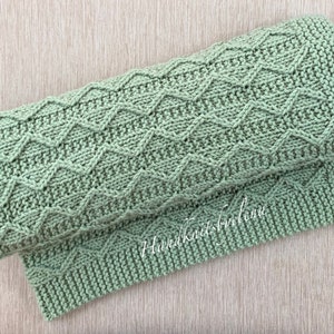 May include: A sage green hand-knitted blanket featuring a textured zigzag chevron pattern across the main body and a garter stitch border. The fabric displays a consistent knit structure with a visible watermark text reading Handknitbyelena.