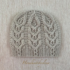 Puede incluir: Un gorro de punto color beige claro con un patrón de trenzas verticales con motivos repetidos en forma de hoja y un borde acanalado. El gorro está confeccionado con un diseño de punto texturizado e incluye el texto Handknitsbyelena abajo.