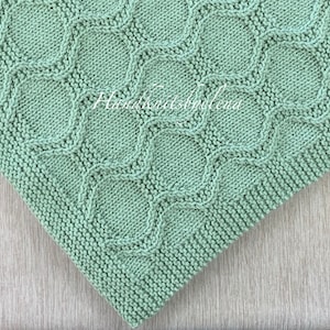 May include: A close-up of a light green knitted blanket with a diamond pattern. The blanket is made with a soft yarn and has a delicate texture.