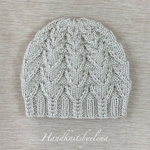 May include: A light gray knit beanie featuring a textured leaf-like cable pattern throughout the body and a ribbed brim at the base. The text Handknitsbyelena is printed in white cursive script at the bottom center of the image.