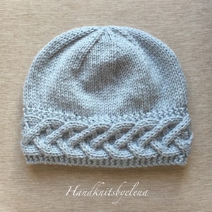 May include: A light gray knitted beanie hat with a braided cable knit design on the brim. The hat is made of soft yarn and features the text "Handknitsbyelena" at the bottom. The hat is a winter accessory.
