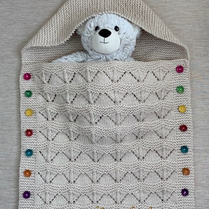 May include: A beige knitted baby blanket with a hood, featuring a geometric pattern and colorful buttons along the side. A plush teddy bear peeks out from the hood. The blanket has the text "HandKnitsbyElena".