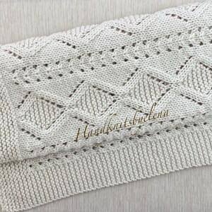 May include: A cream-colored knitted blanket featuring a textured diamond pattern, eyelet details, and a garter stitch border. The text Handknitsbyelena is printed in a gold script font across the center of the folded fabric.