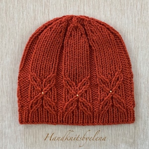 Könnte beinhalten: Eine handgestrickte Mütze in einem tiefen Rostorange mit einem Zopfmuster und goldenen Perlen. Die Mütze hat eine lockere Passform und ist aus weichem Garn gefertigt.