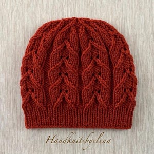 May include: A rust-colored knitted beanie with a textured cable knit design. The hat features a ribbed brim and a detailed pattern across the crown. The text "Handknitsbyelena" is visible at the bottom. This handmade hat is perfect for cold weather.