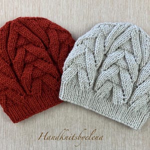 May include: Two knitted beanies. One is a deep red-orange color, the other is a light gray. Both have a textured, braided cable knit design. The beanies are displayed on a light wood-grain surface. The text "Handknitsbyelena" is at the bottom.