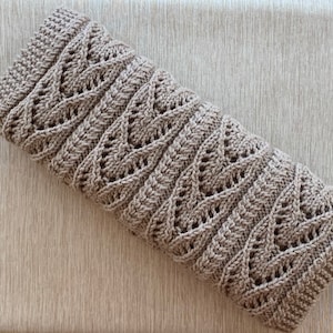 May include: A rectangular, light brown crocheted piece featuring a repeating vertical leaf-like lace pattern. The item is constructed with a dense, textured yarn, showcasing a series of openwork stitches that form distinct, symmetrical foliage motifs.