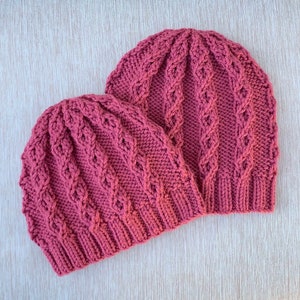 Op de afbeelding: Twee gebreide mutsen in een diepe roze kleur met een kabelbreipatroon.