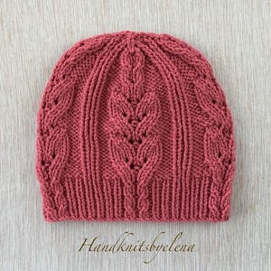 Puede incluir: Un gorro de punto color rosa empolvado con un patrón de trenzas verticales con motivos en forma de hoja y un borde acanalado. El gorro está confeccionado con hilo grueso con un diseño de punto texturizado. El texto Handknitsbyelena es visible abajo.