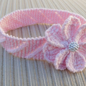 May include: A light pink crochet headband featuring a textured, diagonal ribbed pattern throughout the band. A large, multi-petaled crochet flower is attached to the side, centered with a small, round, silver-toned beaded embellishment.