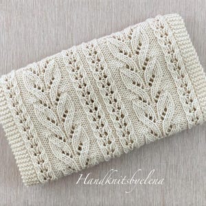 May include: Cream-colored knitted blanket with a detailed cable knit pattern. The rectangular blanket features a textured design with openwork details and a ribbed border. The blanket is folded and lies on a light wooden surface. The text "Handknitsbyelena" is visible.
