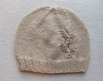 May include: A beige knit beanie featuring a ribbed brim and a delicate leaf-shaped lace pattern on the side. The lace design is accented with small, shiny gold-toned beads integrated into the knit structure, creating a subtle decorative detail.
