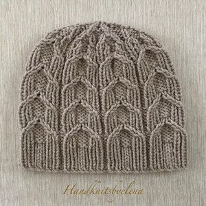 Puede incluir: Un gorro de punto de color marrón claro con un complejo patrón de trenzas repetitivas que crea una serie de estructuras verticales en forma de arco en la superficie. El gorro tiene un borde acanalado y el texto Handknitsbyelena aparece debajo.