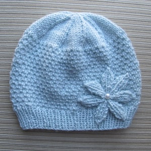 May include: A light blue knitted beanie hat featuring a textured stitch pattern and a matching light blue fabric flower embellishment on the side. The center of the flower is accented with a single small white pearl bead detail.