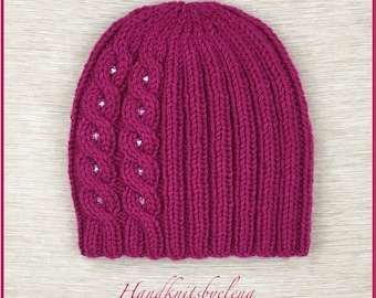 May include: A magenta knit beanie featuring a vertical cable knit pattern on the left side accented with small clear crystals and a ribbed knit texture on the right side. The text Handknitsbyelena is printed in a cursive font at the bottom.