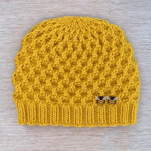 Può includere: Un berretto in maglia color giallo senape caratterizzato da un motivo a nido d'ape testurizzato su tutto il corpo e un bordo a coste alla base. Due piccole decorazioni a forma di ape con strisce nere e gialle sono fissate in basso a destra.
