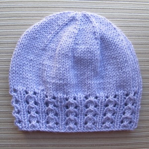 May include: A light lavender knit beanie featuring a smooth stockinette stitch on the crown and a decorative openwork lace pattern along the bottom edge. The hat is constructed from soft yarn and displays a consistent light purple color throughout.