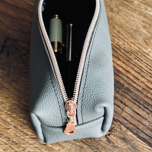 Könnte beinhalten: Ein graues Leder-Etui mit einem roségoldenen Reißverschluss. Das Etui ist geöffnet und enthält einen Stift und einen Bleistift.