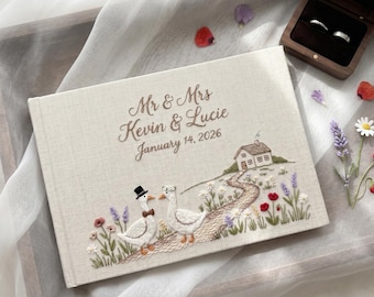Może przedstawiać: Kremowy album ślubny w oprawie z tkaniny z wyhaftowanym tekstem Mr & Mrs Kevin & Lucie January 14, 2026, oraz sceną przedstawiającą dwie gęsi, mały dom i polne kwiaty. Obok znajduje się ciemne drewniane pudełko na obrączki z dwoma srebrnymi pierścionkami.