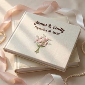 Può includere: Due libri degli ospiti di matrimonio color avorio sono impilati. Il libro superiore è ricamato con i nomi "James & Emily" e la data "14 settembre 2028" con filo marrone, insieme a un bouquet di fiori rosa. Nastri e perle sullo sfondo.