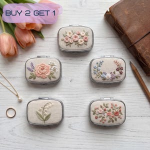 Può includere: Cinque piccoli specchietti compatti rettangolari con disegni floreali ricamati. I compatti hanno cornici in metallo color argento e tessuto beige. Il ricamo presenta fiori rosa, viola e bianchi. Il testo "BUY 2 GET 1" è visibile.