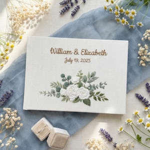 May include: A white fabric-covered guest book featuring embroidered text that reads William & Elizabeth and July 19, 2025, centred above a detailed floral arrangement of white roses and green leaves stitched onto the front cover.