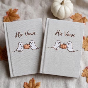 May include: Two light beige hardcover books with embroidered designs. One book reads Her Vows and the other reads His Vows in brown script. Each cover features two white ghosts holding a small orange pumpkin between them with brown thread.